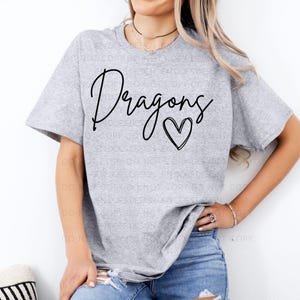 May include: A light gray t-shirt with the word "Dragons" in a black script font and a black heart graphic. The shirt is made of a soft material and has short sleeves.