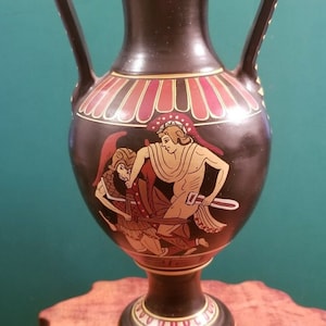 Puede incluir: Jarrón de cerámica negra con dos asas y una escena de dos figuras pintadas en rojo, amarillo y blanco en el lateral. El jarrón tiene un borde dorado alrededor de la parte superior e inferior.