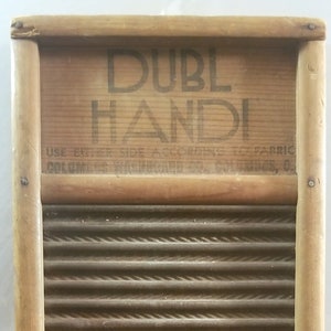 Op de afbeelding: Een houten wasbord met een gegolfd metalen oppervlak. Het wasbord heeft een bord met de tekst "DUBL HANDI" en "USE EITHER SIDE ACCORDING TO FABRIC COLUMBUS WASHBOARD CO. COLUMBUS, O".
