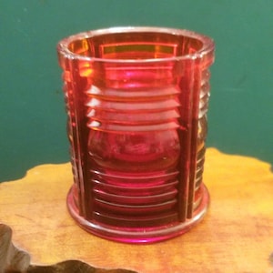 Puede incluir: Un pequeño tarro de vidrio rojo con un diseño acanalado. El tarro tiene un fondo redondeado y una parte superior cilíndrica recta.