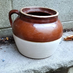May include: A two-tone ceramic crock with a handle. The upper portion is glossy brown, and the lower half is white. The crock has a wide opening and is likely used for storage or decoration. The crock is sitting on a concrete surface.