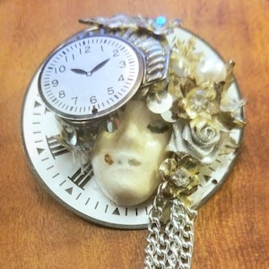 Peut inclure: Un visage en porcelaine blanche avec un cadran d'horloge fixé sur le front. Le cadran de l'horloge a des aiguilles noires et des chiffres romains noirs. Le visage est entouré d'embellissements dorés et argentés, y compris une chaîne argentée.