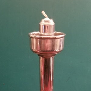 May include: A silver oil lamp with a round base and a tall, cylindrical stem. The lamp has a round, shallow bowl at the top with a wick.