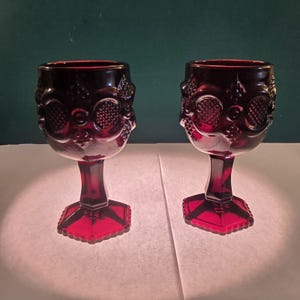 May include: Two ruby red goblets with ornate, textured designs. The goblets have a detailed pattern around the bowl and a faceted base. The stem is a solid, rectangular shape. The background is a dark green.