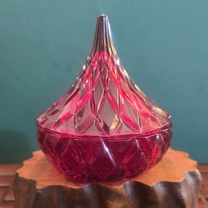 Puede incluir: Un recipiente de cristal de color rojo intenso con un patrón de diamantes facetados y una tapa cónica puntiaguda. El objeto presenta una base redondeada y una superficie translúcida reflectante que capta la luz, creando sombras geométricas.