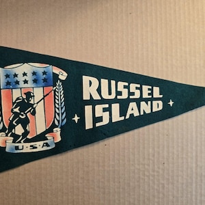 May include: A vintage pennant in teal green with white text reading "RUSSEL ISLAND." The pennant features a shield with a soldier silhouette, stars, and stripes, with "U.S.A." below. A classic souvenir.