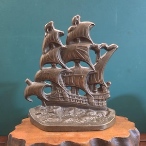 Könnte beinhalten: Bronze-farbene Schiffsskulptur mit detaillierten Segeln und Takelage, montiert auf einem Holzsockel. Das Schiff wird auf einem strukturierten Meer dargestellt, das aufwendige Handwerkskunst zeigt. Ein dekoratives nautisches Objekt.