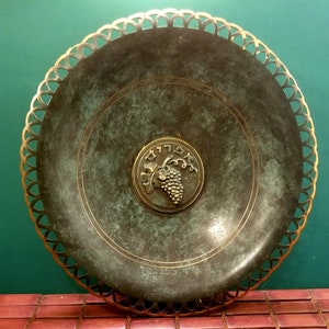 Può includere: Un piatto da portata rotondo in bronzo con un bordo decorativo dorato. Il piatto presenta un design circolare in rilievo al centro con una vite e la parola ebraica "שלום" (Shalom) che significa pace.