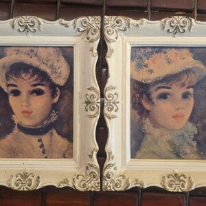 May include: A pair of vintage framed portraits. Each portrait features a woman with dark hair and a hat. The frames are off-white with ornate detailing. The portraits are displayed against a brown background.