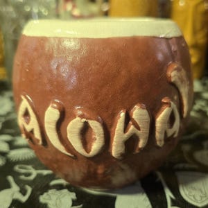 Może przedstawiać: Brązowy ceramiczny kubek z wygrawerowanym słowem "Aloha" z boku. Obręcz jest w kolorze kremowym. Kubek ma zaokrąglony kształt i teksturowaną powierzchnię.