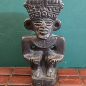 Könnte beinhalten: Eine dunkelgraue, geschnitzte Steinfigur, die eine sitzende Gestalt mit einem markanten, breiten Kopfschmuck mit geometrischen Mustern darstellt. Die Figur hat große, kreisförmige Ohrschmuckelemente, einen breiten Kragen und Hände, die auf den Knien ruhen.