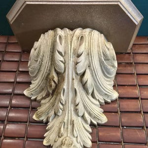 Puede incluir: Soporte de estante blanco adornado con un diseño decorativo en forma de hoja. El soporte de estante tiene una parte superior plana rectangular con un acabado marrón desgastado.