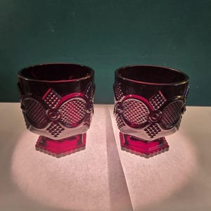 May include: Two ruby red, vintage-style goblets. The glasses have a detailed pattern with diamond and oval shapes. The stem and base are a solid red, while the upper portion is a darker, almost black, red. The goblets are displayed on a white surface.