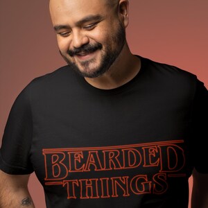 Puede incluir: Camiseta negra con un gráfico rojo que dice "Bearded Things" en un estilo que recuerda al logotipo de Stranger Things.