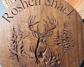 May include: A wooden cribbage board with a carved deer scene and the words "Roshell Shack". A deck of Bee playing cards is visible in the background. The board is made of a dark wood.