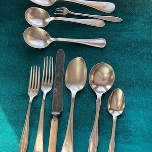 May include: A collection of antique silverware, including spoons, forks, and a knife, arranged on a dark green surface. The silverware is silver-toned, with some pieces bearing engraved initials. The knife has a cream-colored handle.