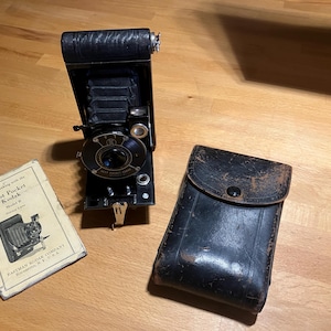 May include: A vintage black folding camera with a leather case and instructions for the Vest Pocket Kodak Model B. The camera has a lens with a silver rim and a black leather strap.