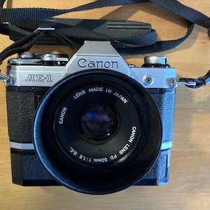 Puede incluir: Una cámara de película Canon AE-1 plateada con un objetivo Canon FD 50mm 1:1.8 S.C. negro. La cámara tiene una correa de cuero negra.