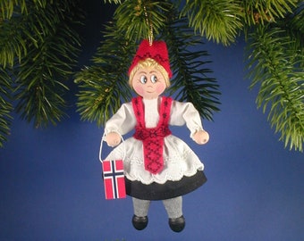 May include: A handmade Christmas ornament depicting a girl wearing a red and white traditional Norwegian folk costume, holding a small Norwegian flag. The ornament is hanging from a pine tree branch.