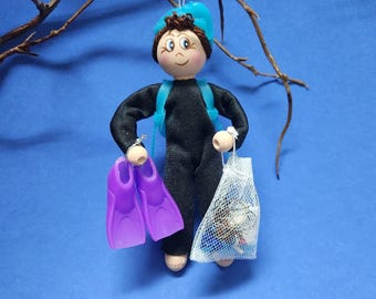 May include: A handmade ornament of a person wearing a black wetsuit, a blue hat, and holding purple flippers and a mesh bag filled with white and blue objects.