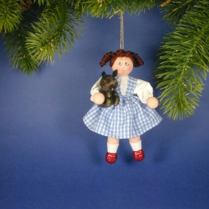 May include: A handmade ornament featuring a girl doll with brown hair, a blue and white gingham dress, and red shoes. She is holding a small brown and black dog.