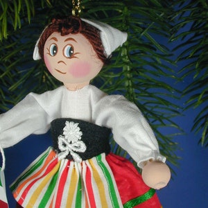May include: A handmade wooden doll ornament with a white bonnet, white blouse, black belt, and a red, yellow, and green striped skirt. The doll has brown hair and painted features.