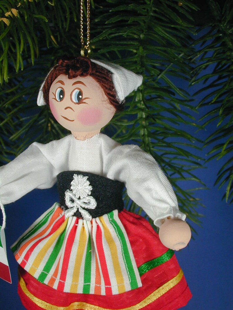 May include: A handmade wooden doll ornament with a white bonnet, white blouse, black belt, and a red, yellow, and green striped skirt. The doll has brown hair and painted features.