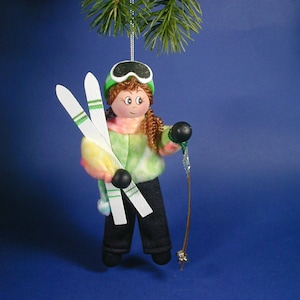 May include: A handcrafted Christmas ornament depicting a skier. The figure wears a green hat and goggles, a colorful sweater, and black pants. The ornament holds skis and ski poles, and is suspended from a pine branch.