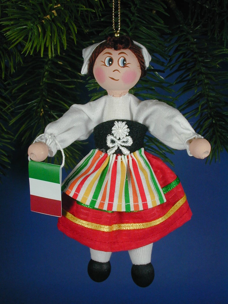 May include: A handmade wooden doll ornament dressed in a traditional Italian costume. The doll is wearing a white blouse, a green and red striped skirt, and a red apron. The doll is holding a small Italian flag.