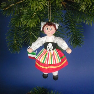 May include: A handmade ornament depicting a girl in traditional Italian clothing. She is wearing a white blouse, a red and green striped skirt, and a red apron. She is holding a small Italian flag. The ornament is made of fabric and is hanging from a gold string.