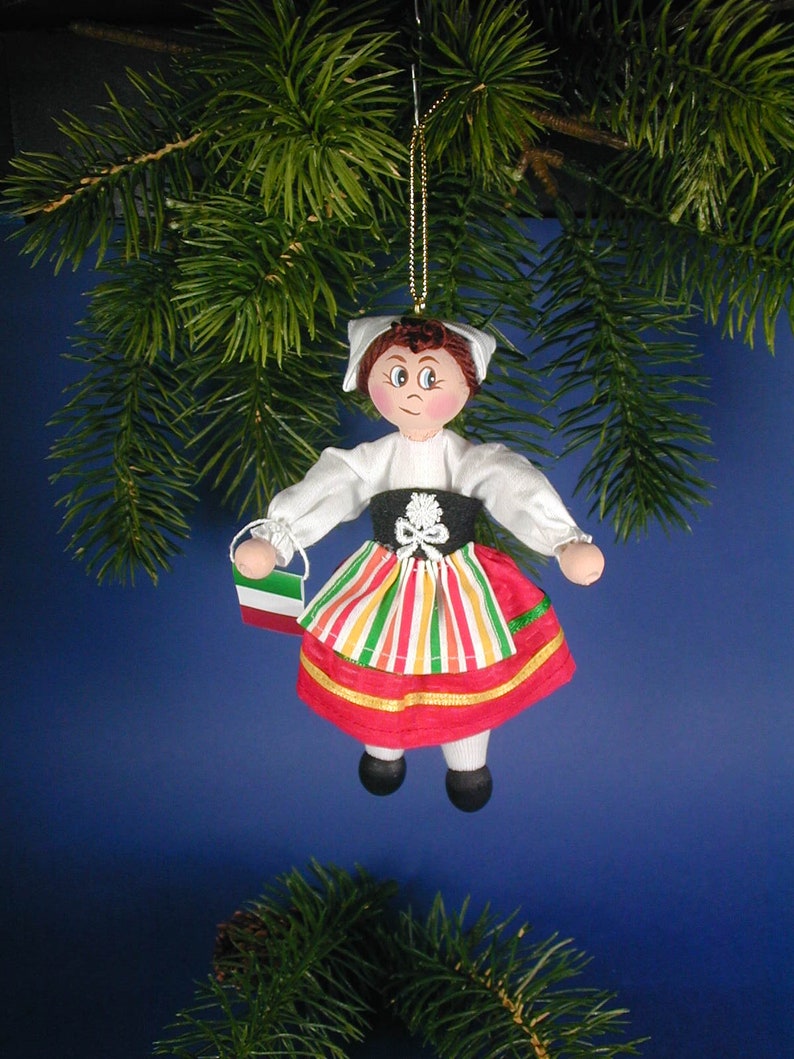 May include: A handmade ornament depicting a girl in traditional Italian clothing. She is wearing a white blouse, a red and green striped skirt, and a red apron. She is holding a small Italian flag. The ornament is made of fabric and is hanging from a gold string.