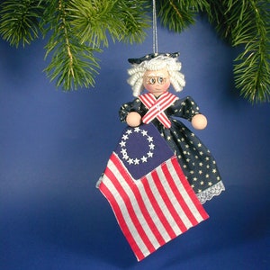 May include: A handmade doll ornament dressed in a blue and white dress with a red sash and a white lace trim. The doll is holding a small American flag with 13 stars.