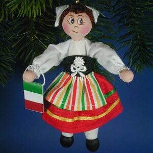 May include: A handmade doll ornament wearing a traditional Italian dress with a red, green, and white striped skirt and a white blouse. The doll is holding a small Italian flag.