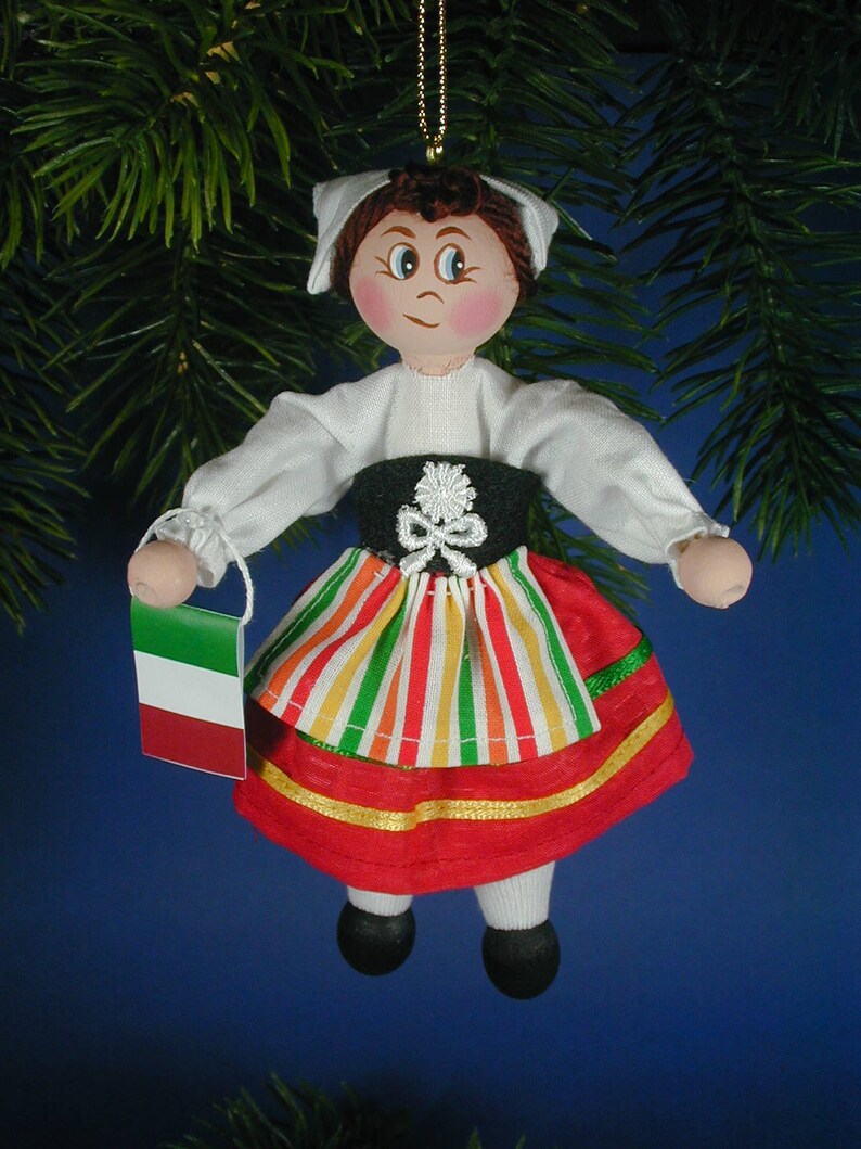 May include: A handmade doll ornament wearing a traditional Italian dress with a red, green, and white striped skirt and a white blouse. The doll is holding a small Italian flag.