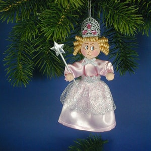 May include: A pink and silver fairy doll ornament with a crown and a wand. The doll has blonde hair and a painted face. The ornament is hanging from a tree branch.