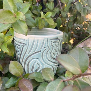 May include: A handcrafted ceramic mug with a pale blue glaze and a textured, carved pattern. The mug is set amongst green leaves, giving a natural, earthy feel. The rim is a lighter shade of white.