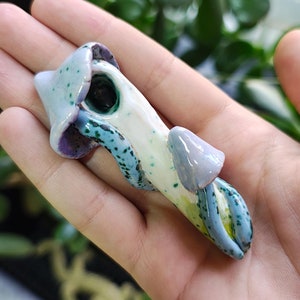 May include: A ceramic pendant shaped like a mushroom with a blue and green glaze. The mushroom has a white body and a purple cap.