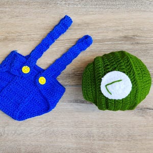May include: A handmade crochet outfit, featuring a blue romper with yellow buttons and suspenders, and a green hat with a white circle and a green embroidered detail. The set is designed for a baby.