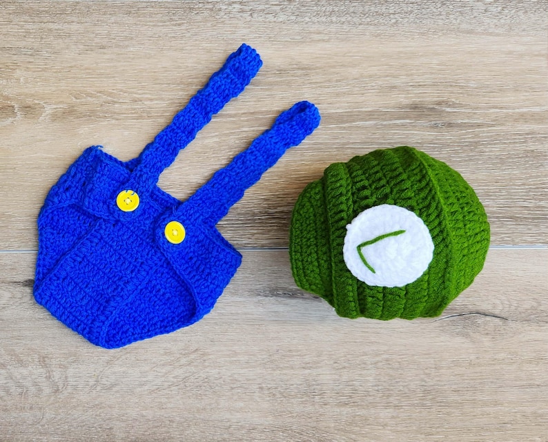 May include: A handmade crochet outfit, featuring a blue romper with yellow buttons and suspenders, and a green hat with a white circle and a green embroidered detail. The set is designed for a baby.