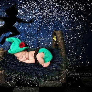 May include: A sleeping baby wearing a green crocheted hat with a red flower and matching green diaper cover. The baby is lying on a dark blue surface with a Peter Pan silhouette and fairy dust in the background.