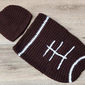 May include: A dark brown crocheted football-themed baby outfit, including a beanie and a cocoon. The cocoon features white stitching details and laces, resembling a football. The set is designed for newborns.