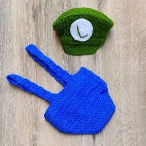 May include: A handmade crochet baby outfit in green and blue. The green hat features a white circle with a green "L". The blue suspenders are connected to a blue hexagon-shaped bottom. The outfit is designed for a baby.