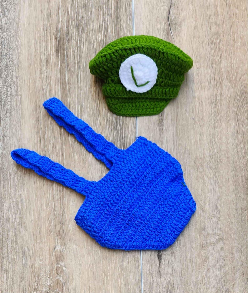 May include: A handmade crochet baby outfit in green and blue. The green hat features a white circle with a green "L". The blue suspenders are connected to a blue hexagon-shaped bottom. The outfit is designed for a baby.