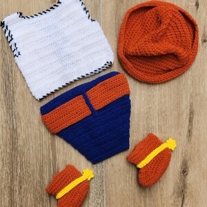 May include: A handmade crochet baby outfit set. The set includes a white vest with black trim, a blue nappy cover with orange accents, an orange beanie, and two orange booties with yellow accents. The items are on a wooden surface.