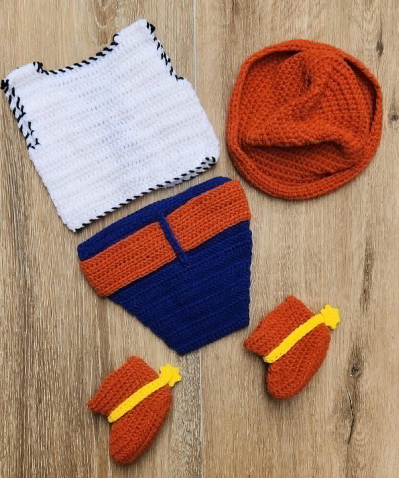 May include: A handmade crochet baby outfit set. The set includes a white vest with black trim, a blue nappy cover with orange accents, an orange beanie, and two orange booties with yellow accents. The items are on a wooden surface.