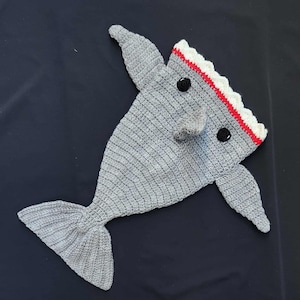 May include: A crocheted shark-shaped baby blanket in gray. The blanket features fins, a tail, and a head with black button eyes and a mouth lined with red and white. The blanket is designed to resemble a shark.