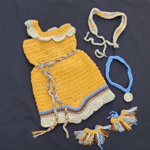 Op de afbeelding: Een handgemaakt gehaakt poppenoutfit in geel, blauw, bruin en crème. De set bevat een jurk, een hoofdband, een ketting en twee oorbellen. De jurk heeft een geschulpte halslijn en zoom en een gevlochten riem. De accessoires zijn ook gehaakt.