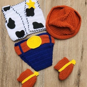 May include: A handmade crochet baby outfit in a cowboy theme. The set includes a white vest with black cow spots, an orange hat, blue and orange diaper cover, and orange booties with yellow stars.