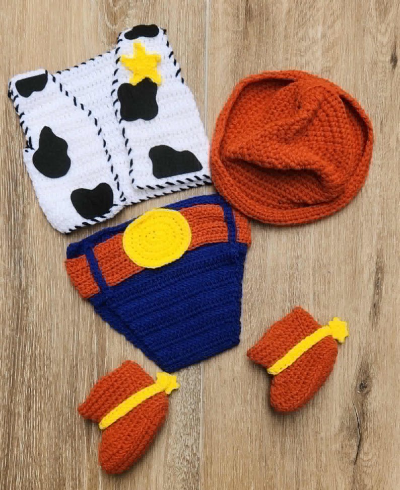 May include: A handmade crochet baby outfit in a cowboy theme. The set includes a white vest with black cow spots, an orange hat, blue and orange diaper cover, and orange booties with yellow stars.