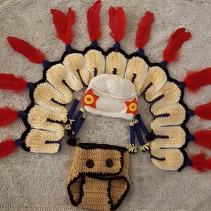 May include: A crocheted baby outfit, featuring a headdress with red feathers, a small white hat with star accents, and a diaper cover. The set is primarily cream-colored with blue and black trim.